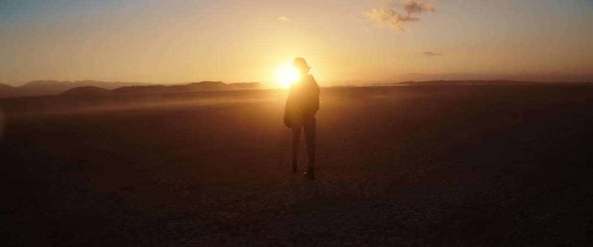 Wind blows sand as silhouetted person walks across desert toward sun sitting on red horizon over mountains