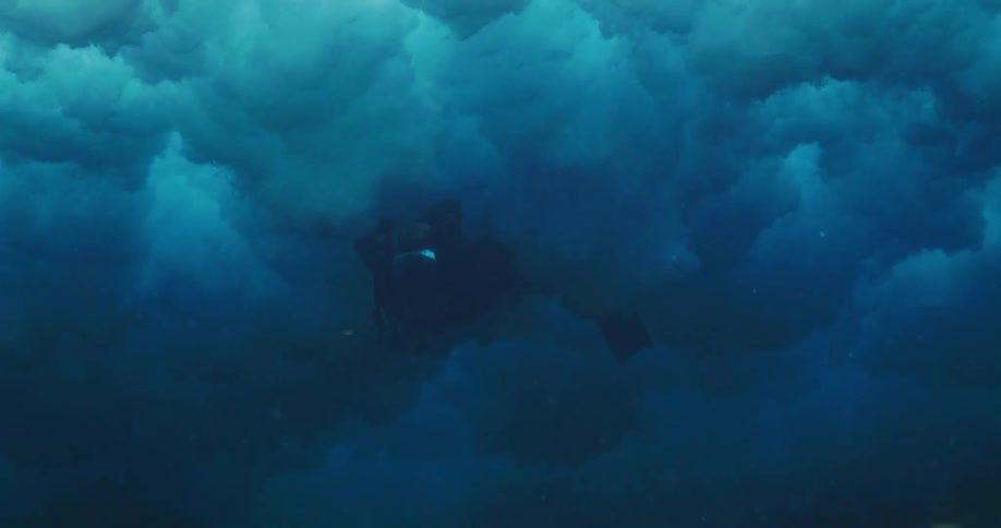 Scuba diver aims underwater camera while swimming above corral as crashing wave flows over area