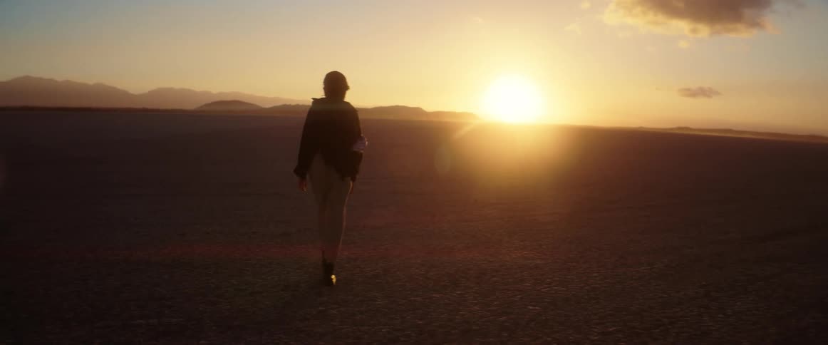 A woman is walking across the desert and the sun is shining brightly on the horizon and there are mountains in the distance