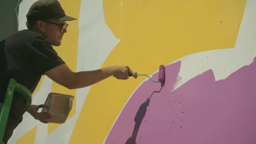 A man uses a paint roller to paint a wall while standing on a raised platform outdoors during the day