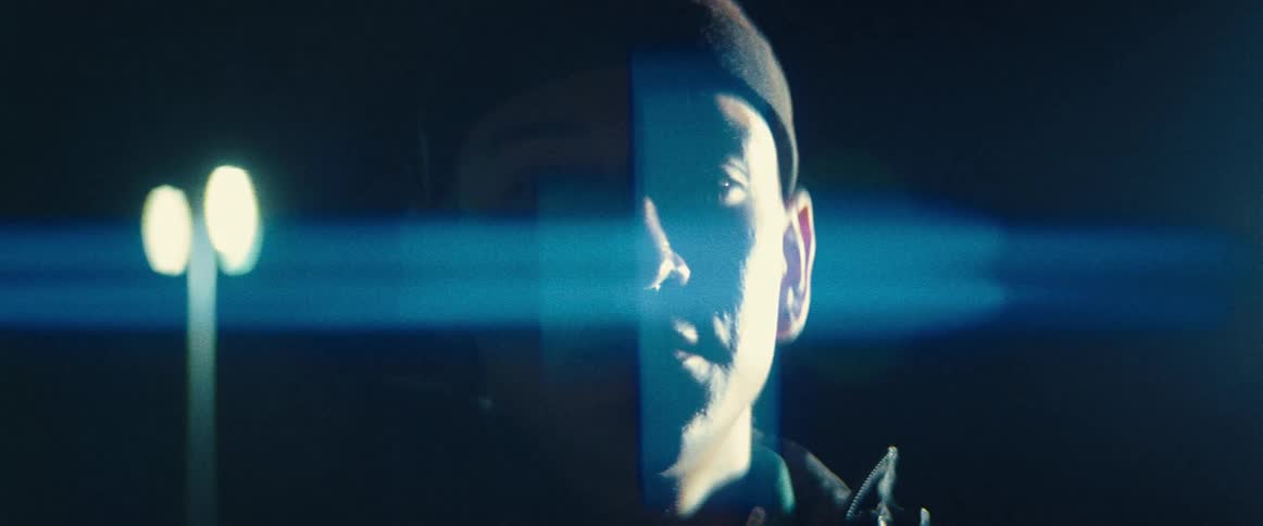 Blue colored light shines on half of mans face leaving the rest in shadow as he stands outdoors at night