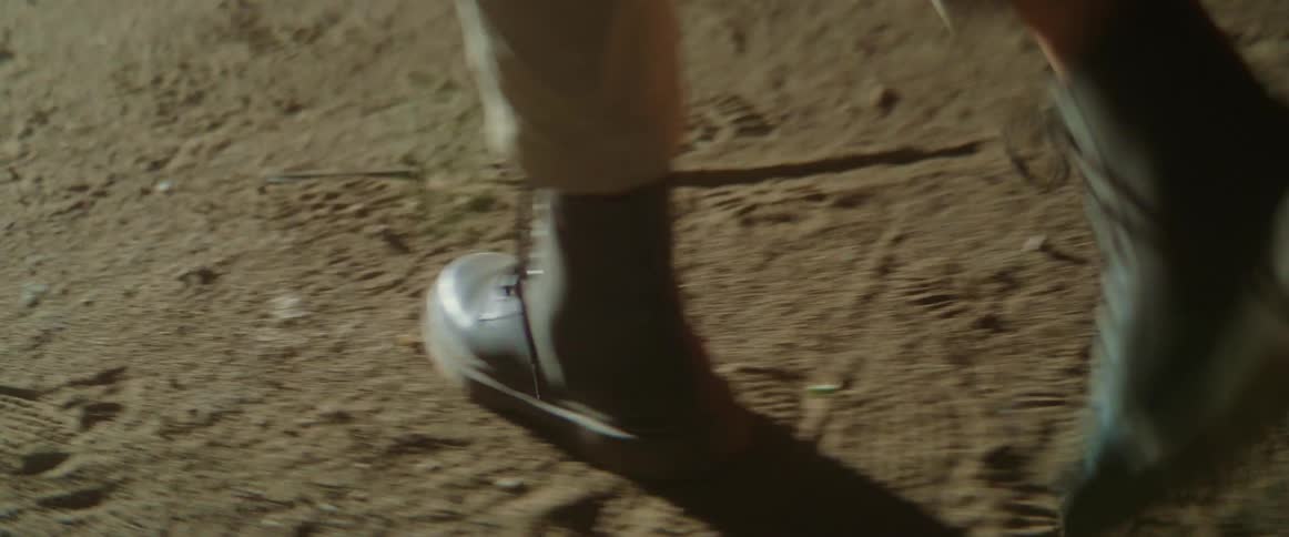 A person wearing lace up boots walks across the dirt and sand from right to left