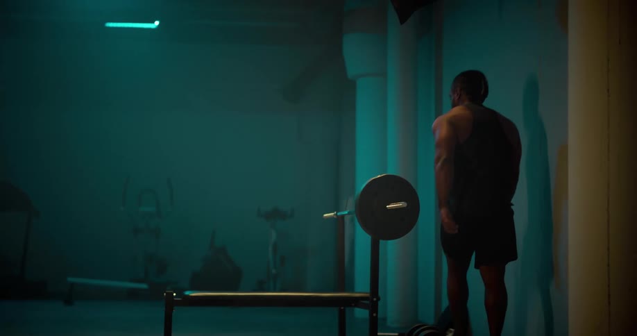 A muscular man approaches a weight bench and attaches weights to the barbell mounted there
