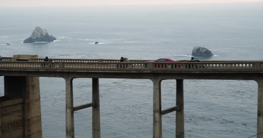 A group of motorcyclists ride down a bridge over the ocean