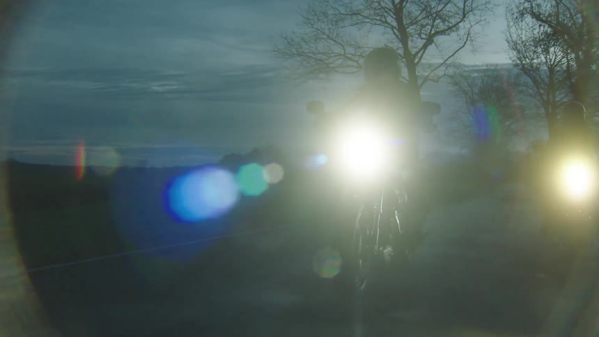 Two cyclists are riding motorcycles on a rural road lined with trees as it grows dark outside