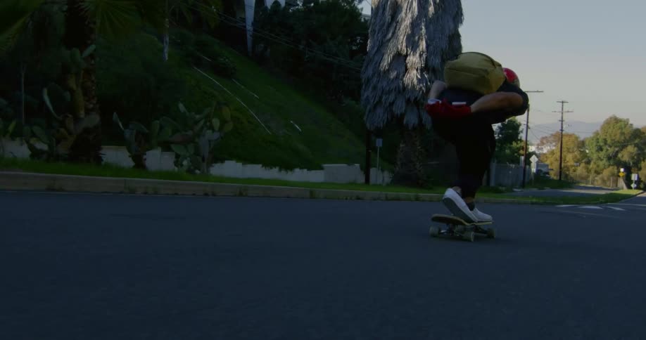 A teenage boy wearing a backpack is coasting downhill on his skateboard on a neighborhood street