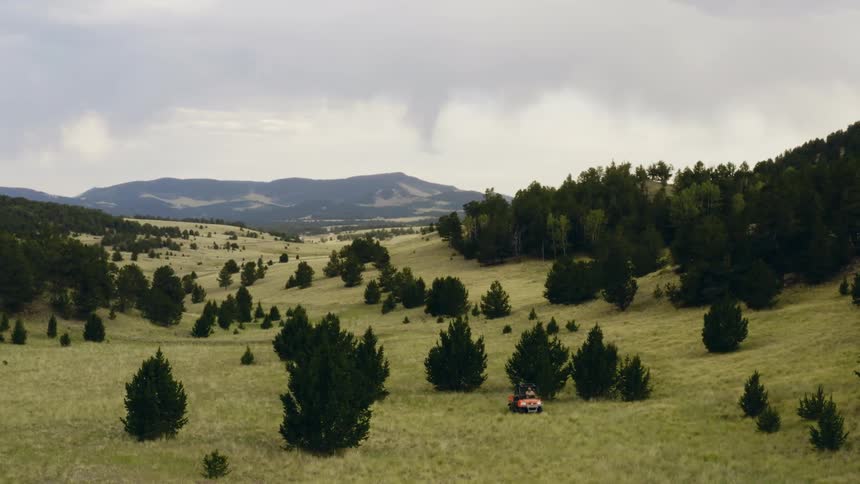 Aerial view of a vehicle driving on the tall green grass in the hills near the mountains