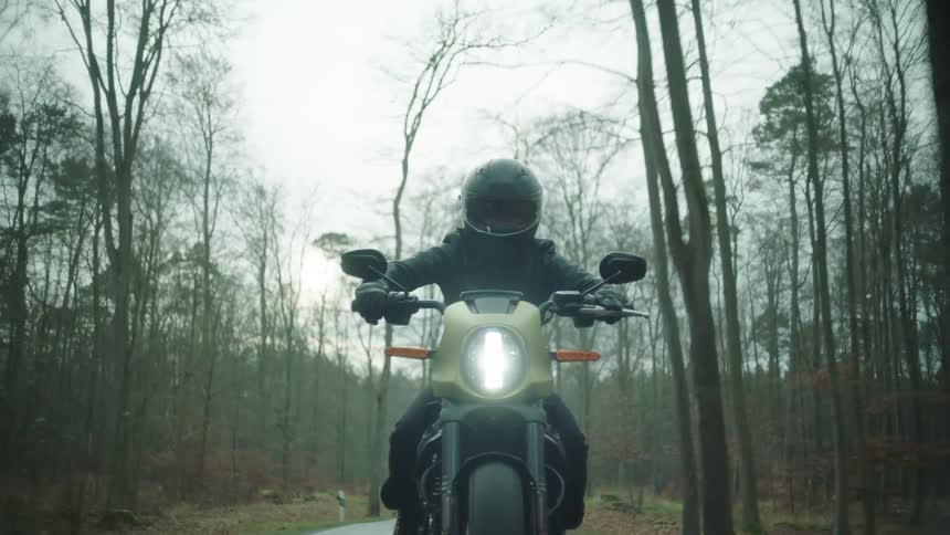 A cyclist wearing a black leather jacket and a helmet is riding a motorcycle on a road through the woods