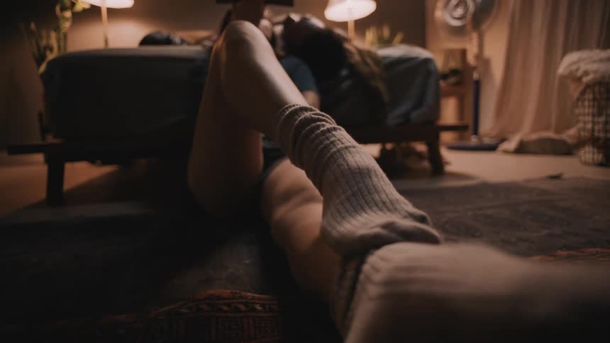 Woman rubs one sock covered foot up her leg while sitting on floor in bedroom reading