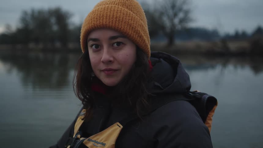 A young woman holding a paddle smiles into the camera while sitting in a boat on a lake on a cloudy day