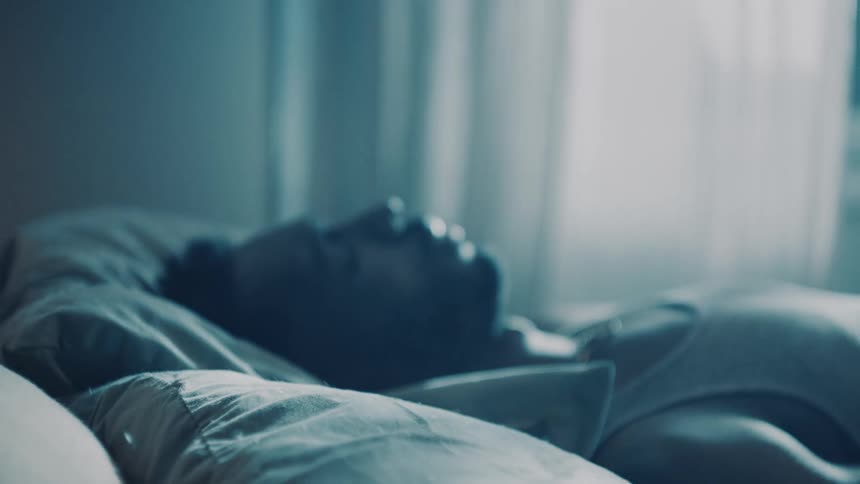 Man collapses back onto bed in dim bedroom