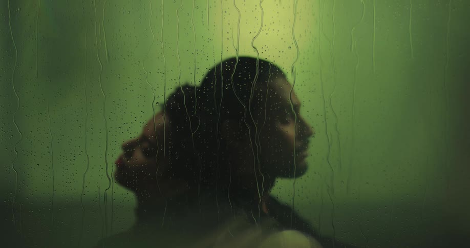 Blurry silhouette of a couple sitting reclined on each others back behind a green wet glass