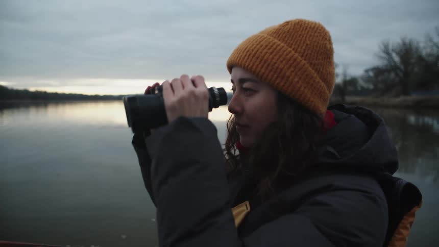 Woman standing near water bordered by forest scans area with binoculars