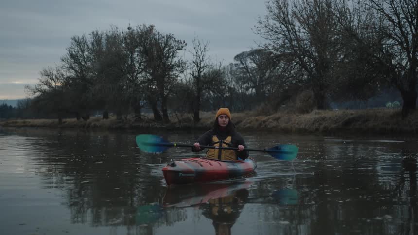 A young woman paddles a kayak across a lake on a cloudy evening