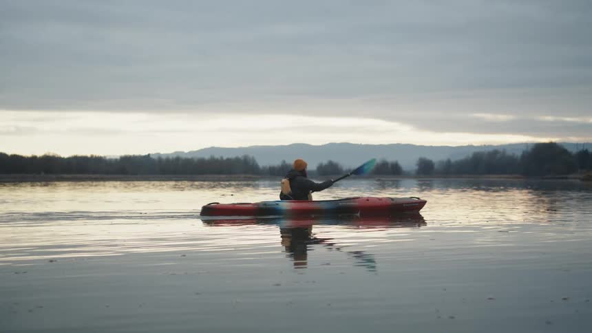 Woman paddles boat toward shore of leafless trees with foggy forest and mountains in the background
