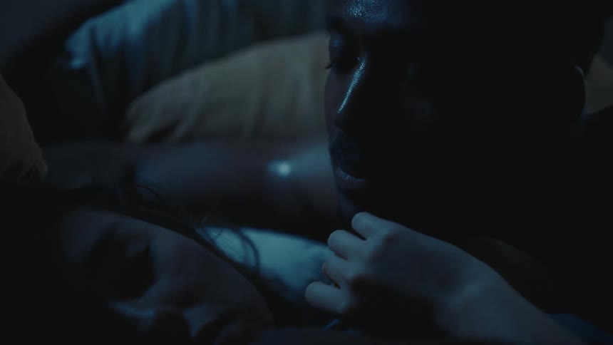 A man leans over a woman in bed and talks to her and she reaches over and covers his mouth and shakes her head and puts her finger over his mouth
