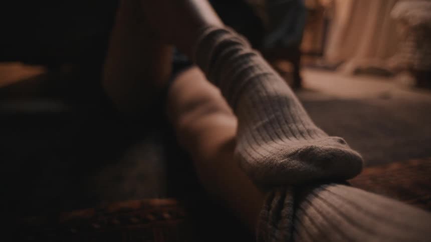 A woman wearing socks is sitting on the floor and caressing her leg with her foot