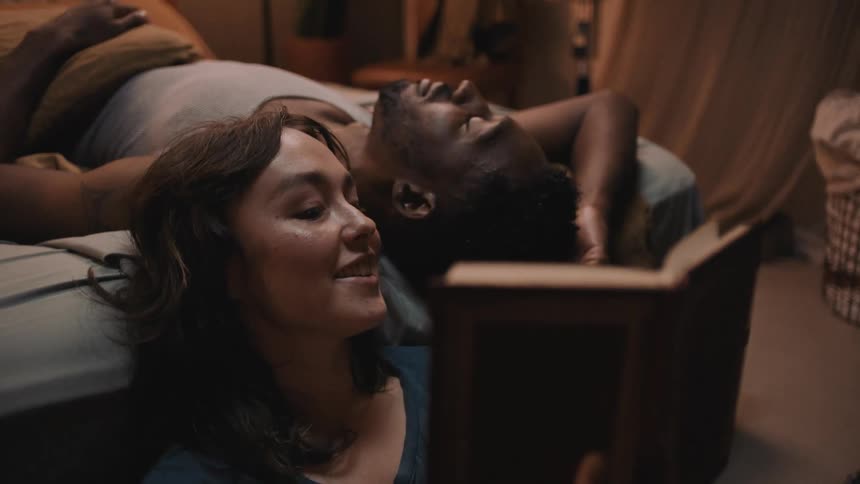 Woman sits on floor reading a book out loud to man lying on bed