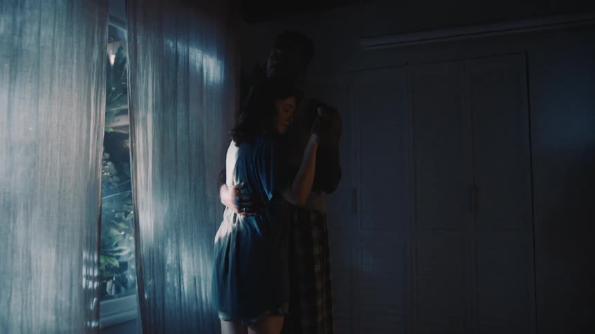 A man and woman are dancing close in a living room by a window at night