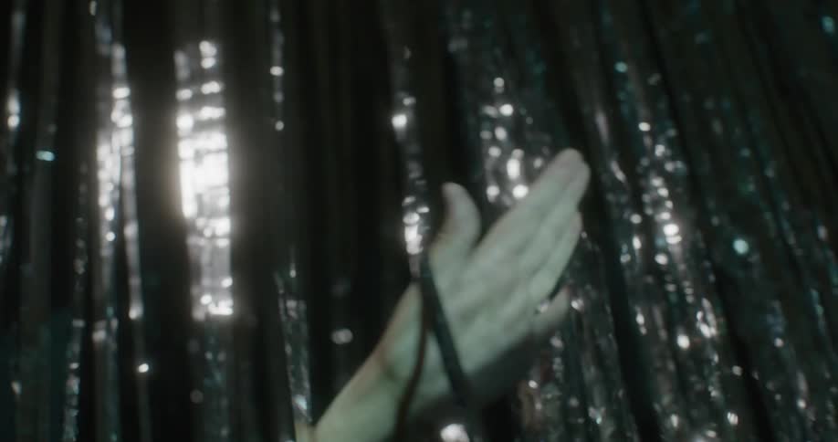Light shines and reflects from silver metalic strips hanging from the ceiling as a woman moves her hands through them