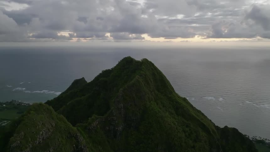 Plants and trees grow on mountains with peaks high in a cloudy sky as waves roll over the water in the distance