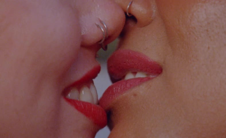 Two women wearing lipstick and nose rings kiss and smile