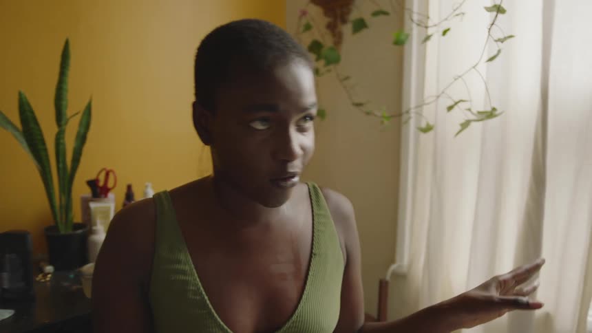 Woman stands near curtained window with table in background and gestures as she talks