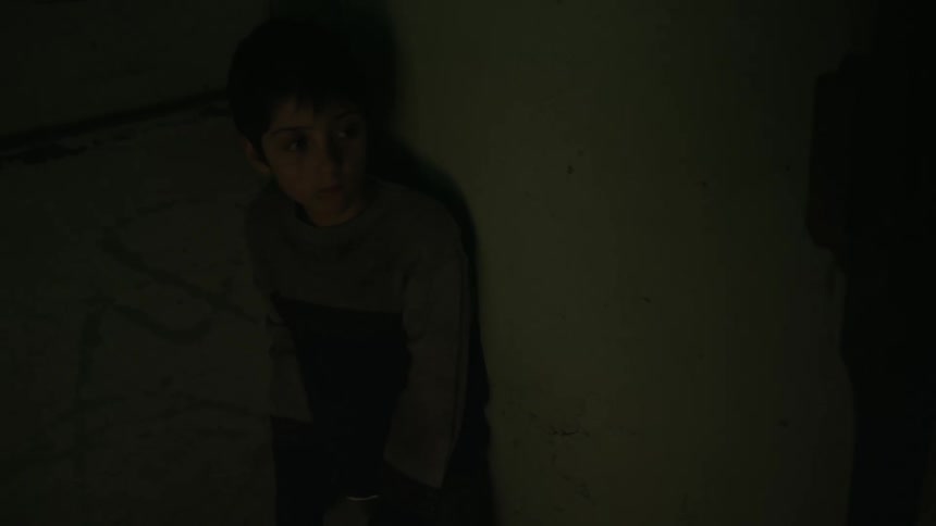 A boy cowers against a wall in a dark room