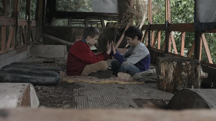 Two boys sit cross legged facing each other in trailer with hands almost touching