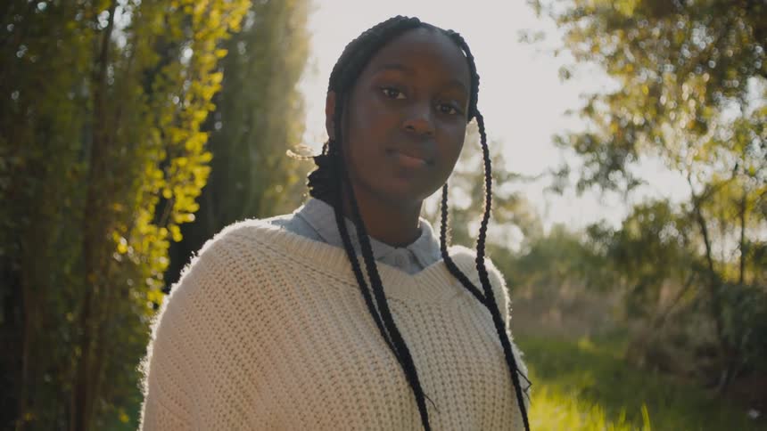 A teenage girl wearing a sweater over her shirt is standing outside with trees in the distance
