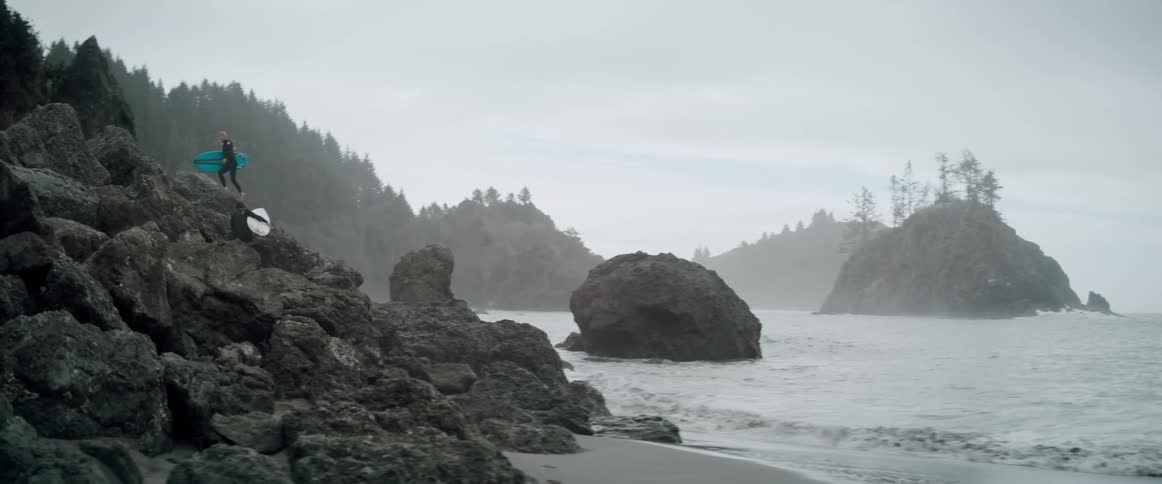 Two men are carrying their surfboards up a rock covered hill as waves splash agains rocks along the shoreline