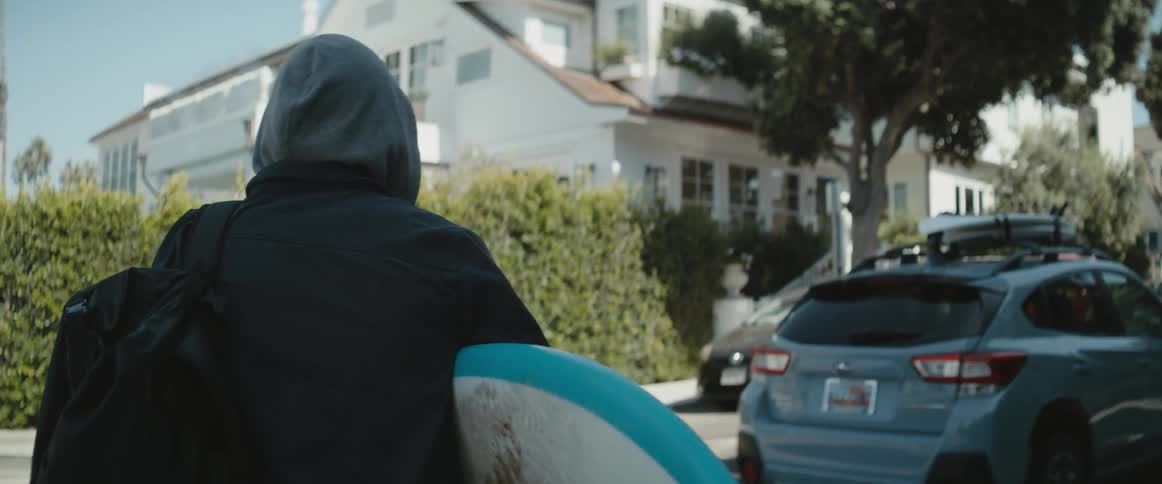 Man holding a surfboard waits for a car on the neighborhood street at morning