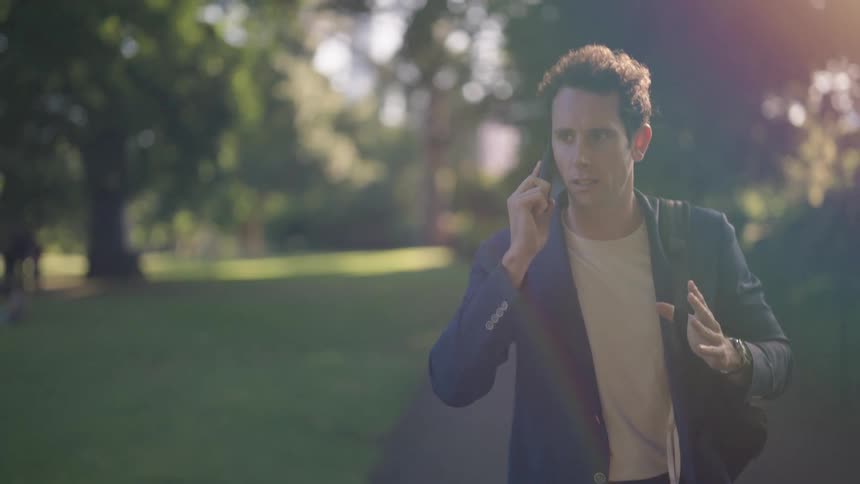 A man carrying a shoulder bag is talking on the phone as he walks through a park