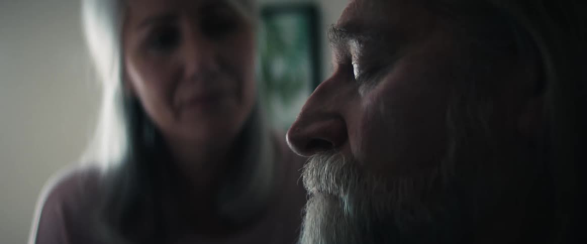 Older woman frowns then leans forward to kiss older man siting with eyes closed