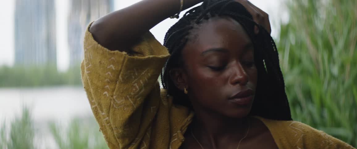 Woman with braids earrings and yellow shirt touches her hair next to the river