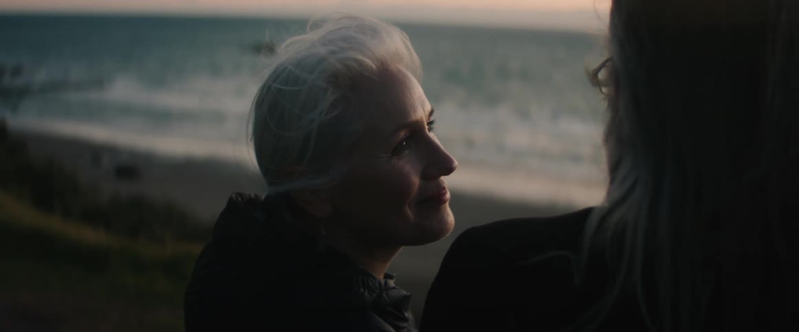 Woman with gray hair and black coat approaches man with long gray hair beard glasses and black coat looking into each others eyes lovingly on the cliff overlooking beach at sunset