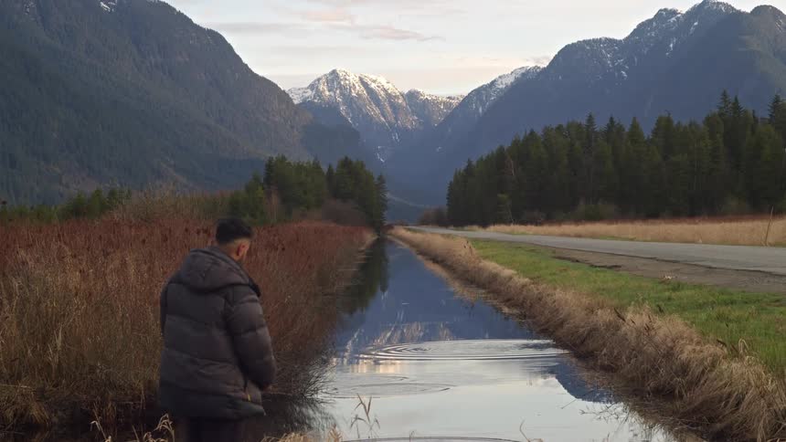 Man skips rocks in stream paralleling highway leading past conifer forest and toward mountains