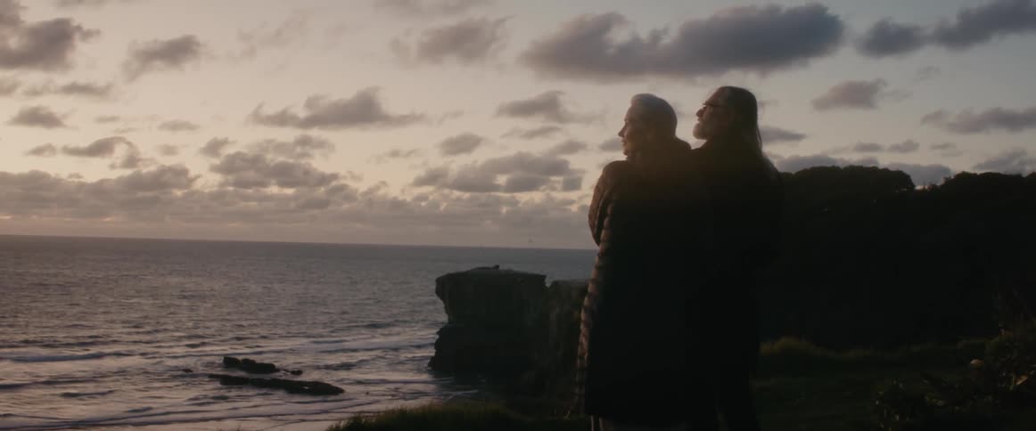 A woman standing on the beach raises her head from a mans shoulder as they look out at the water