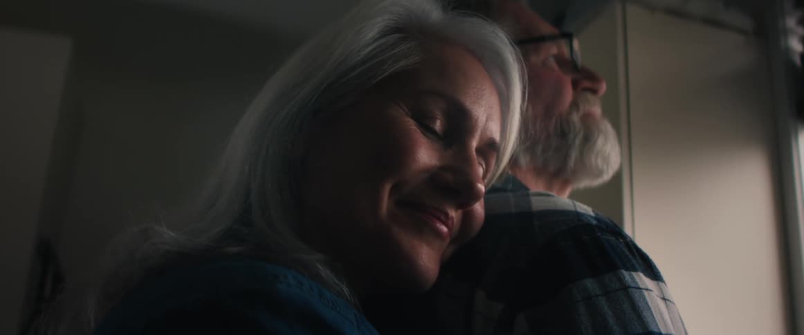 A woman hugs a man from behind and rests her head on his shoulder as he gazes out of a window