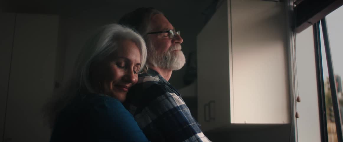 A man is standing in front of a window and his wife is behind him with her arms around his waist and her head on his shoulder