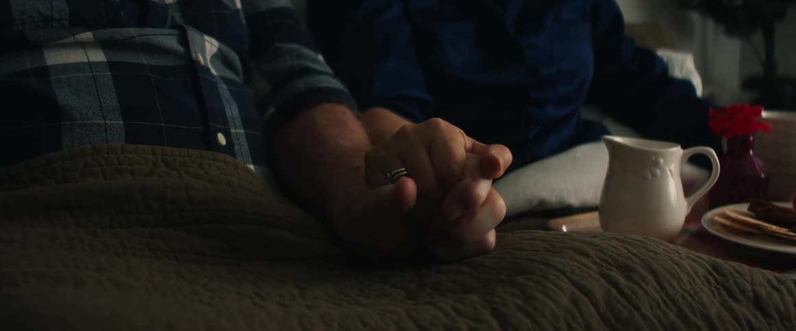Couple holds hands while sitting under blankets with breakfast