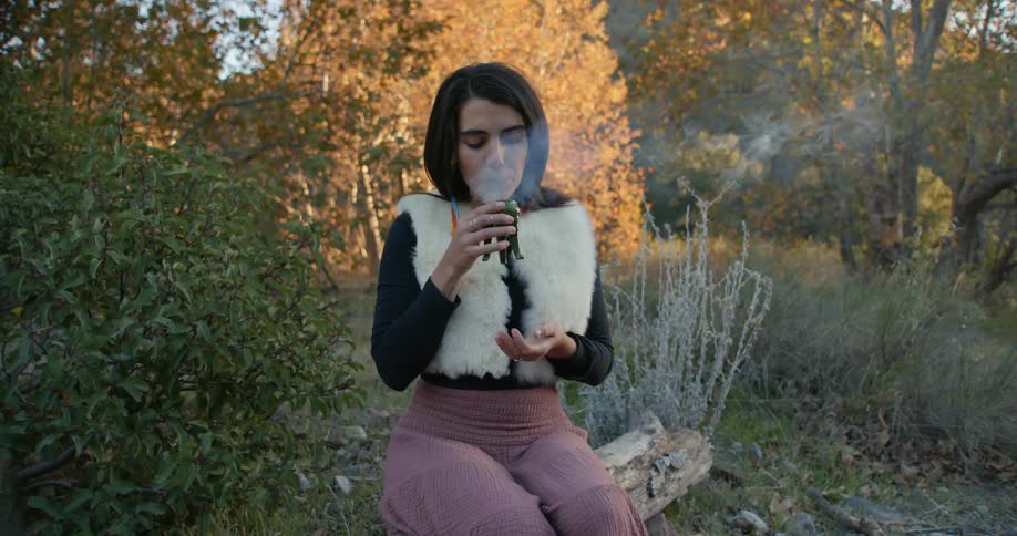 Woman sitting over a log outdoors holds a censer burning aromatic incense at evening