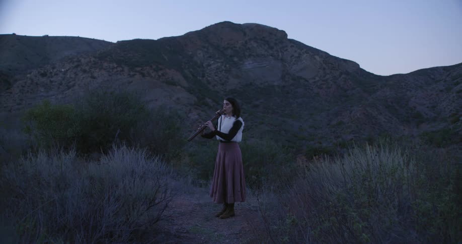 Woman wearing a long skirt plays a long wooden flute on a valley in the mountains at morning