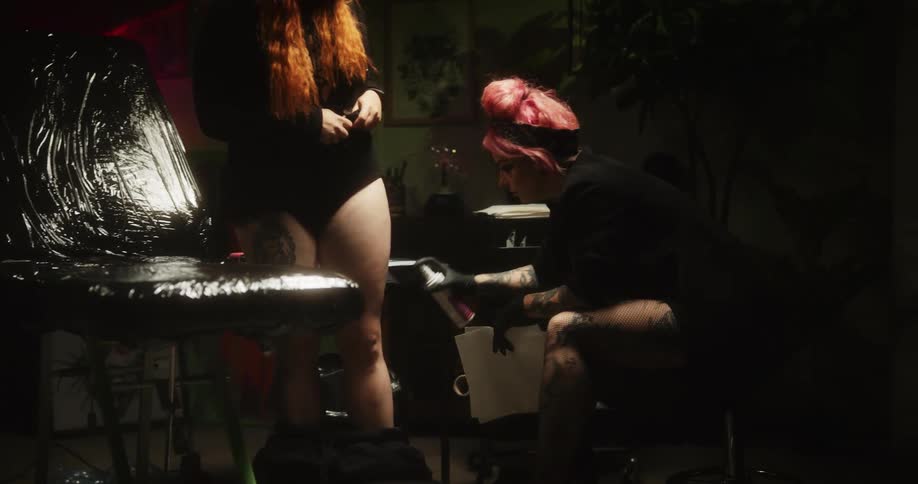 A woman in a tattoo parlor looks at another womans leg and sprays it