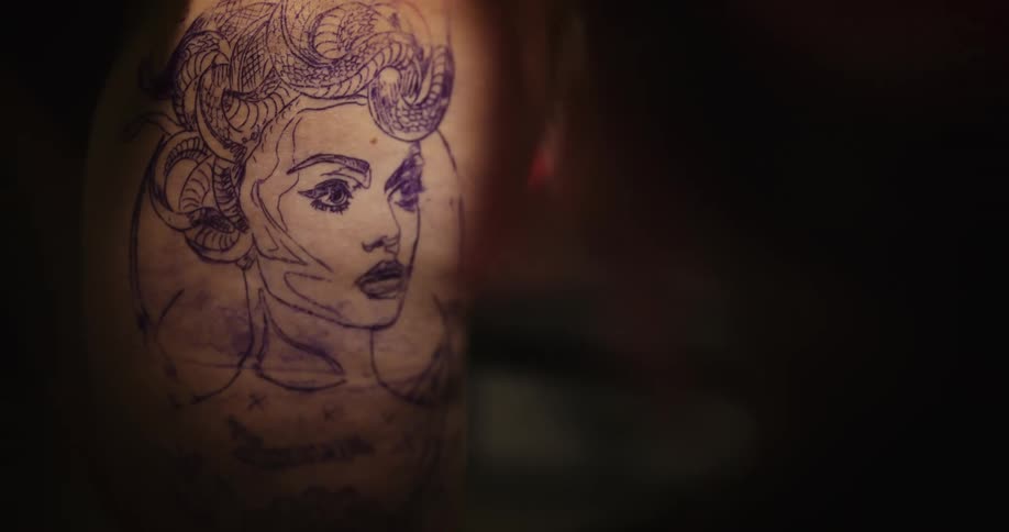 Tattoo artist uses a drawing on paper as a guide to sketch over the client skin