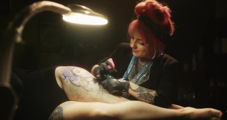 A woman is laying down on her side exposing her thigh where another woman is tattooing her