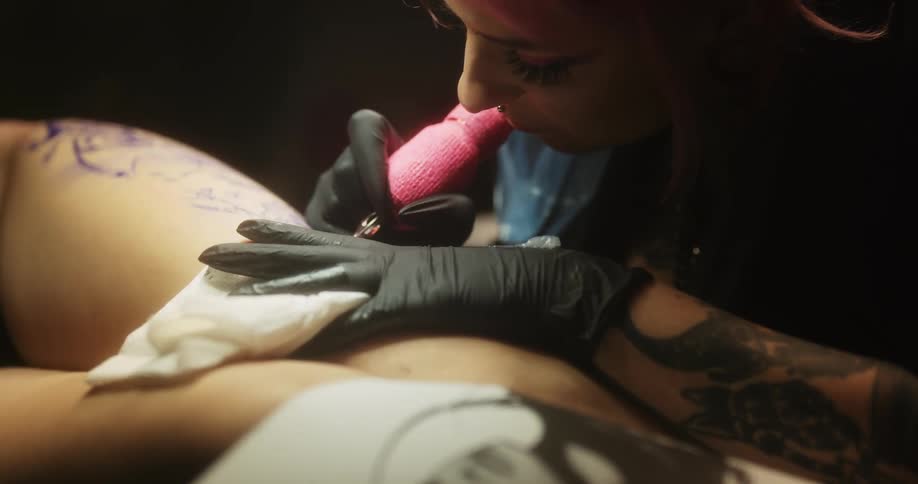 A tattoo artist leans over a womans thigh and applies ink as she tattoos a photo that is lying on the woman