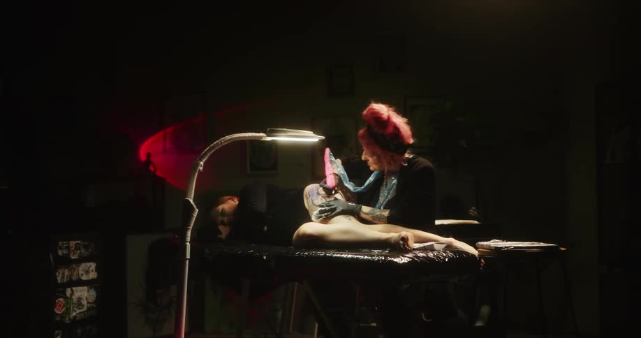 Tattoo artist with colorful hair working on the skin of a client laying on a table