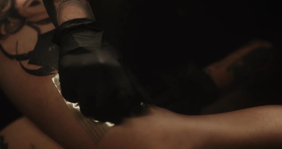 A tattoo artist wearing gloves puts a clear tape bandage on a womans new tattoo on her leg