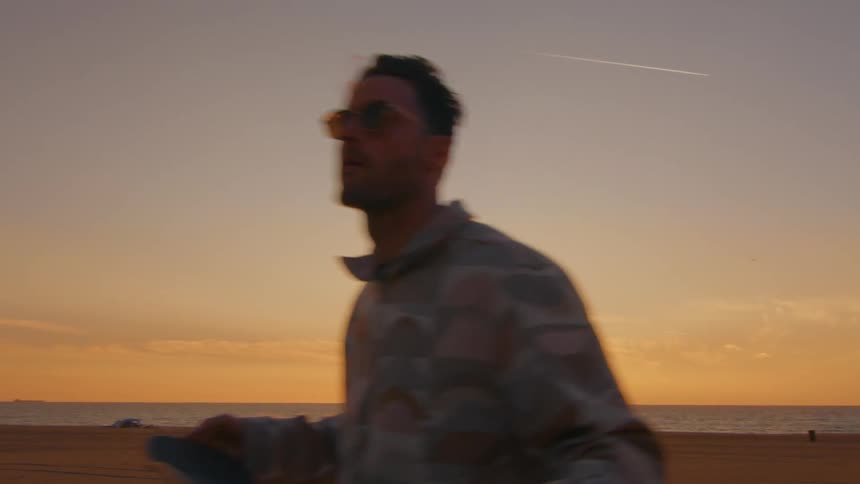 A man is walking and carrying his skateboard on a road along the beach as the sun is setting on the horizon